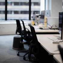 A row of modern, unoccupied office workstations with computers and chairs, set against a backdrop of large windows overlooking a blurred city building.